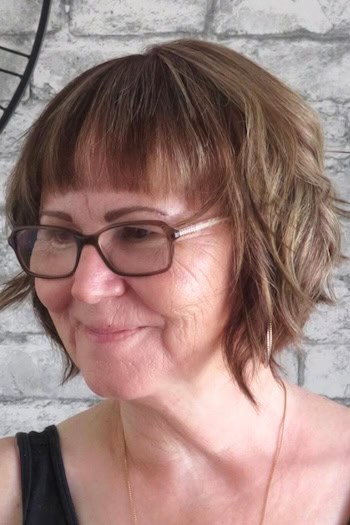 Wavy Layered Bob With Blunt Bangs Hairstyle on a woman in her 60s wearing glasses.