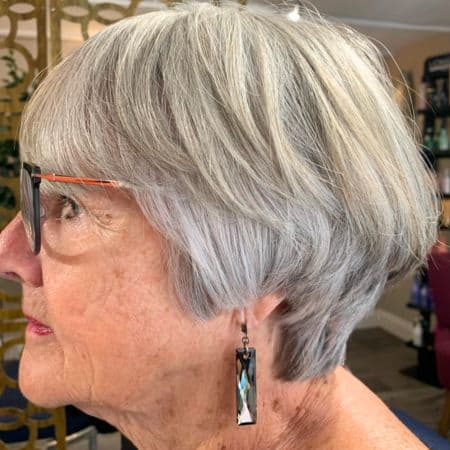 Layered Gray Bob With Bangs Hairstyle on a woman in her 60s wearing glasses, side view.