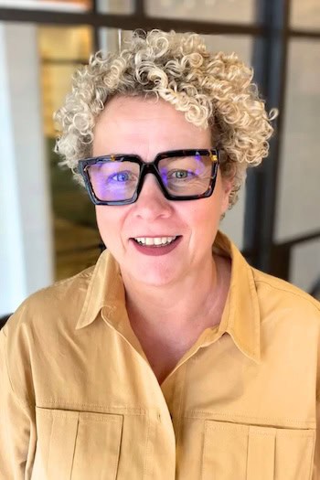 Curly Blonde Pixie Hairstyle, on smiling woman in her 50s with blonde hair and large framed glasses.